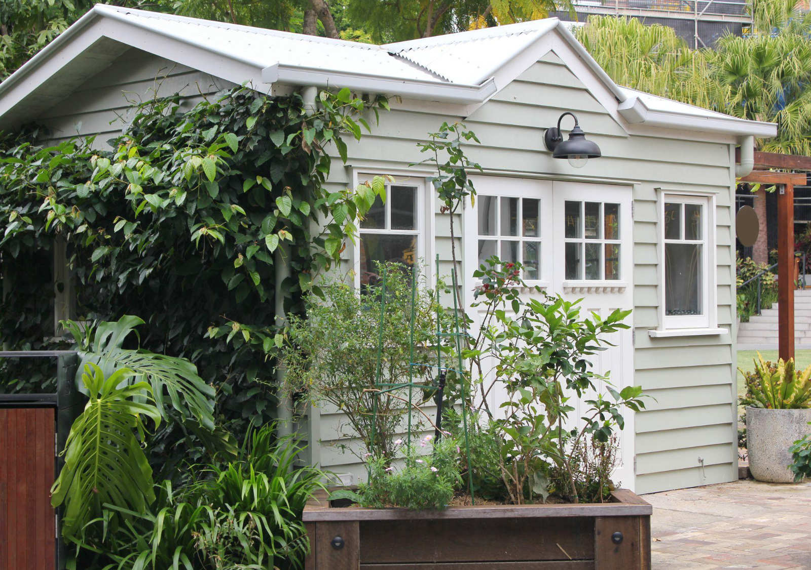 A light green backyard garden shed with white trim and a black gooseneck exterior light fixture