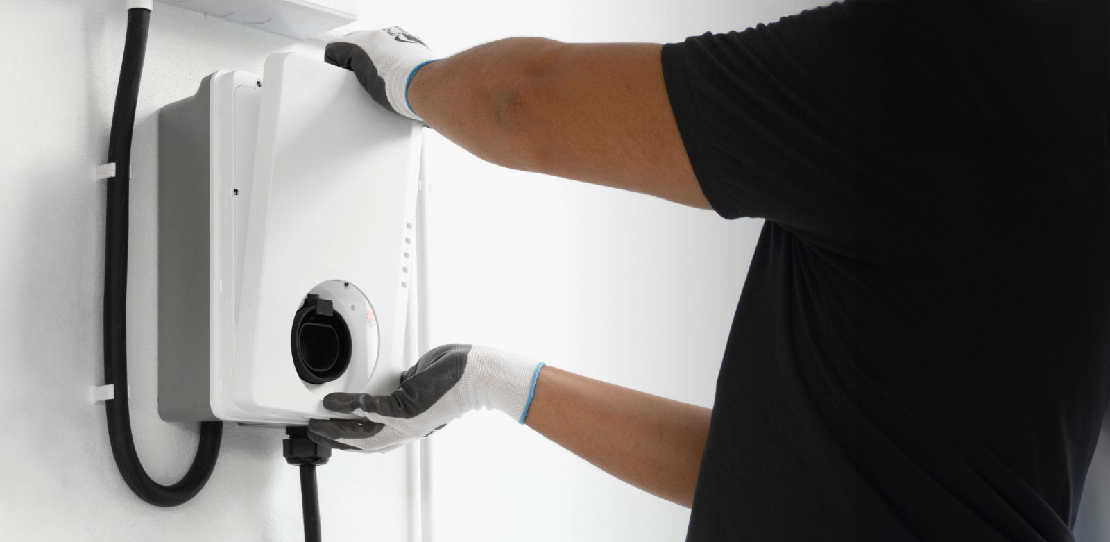 An electrician wearing protective gloves installs a white Level 2 EV charging station on a residential wall.