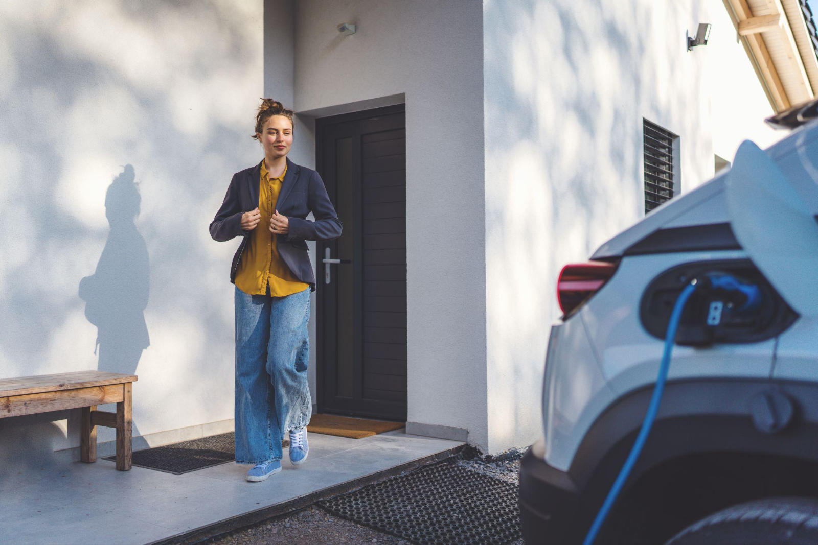 A woman stepping outside her home next to a white electric car plugged into a home wall charger.